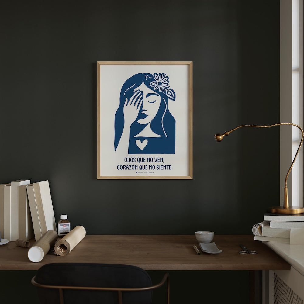 a framed poster of a woman with a flower in her hair, hanging on a dark wall above a wooden desk. The desk has various items on it, including a lamp, books, and a roll of tape.