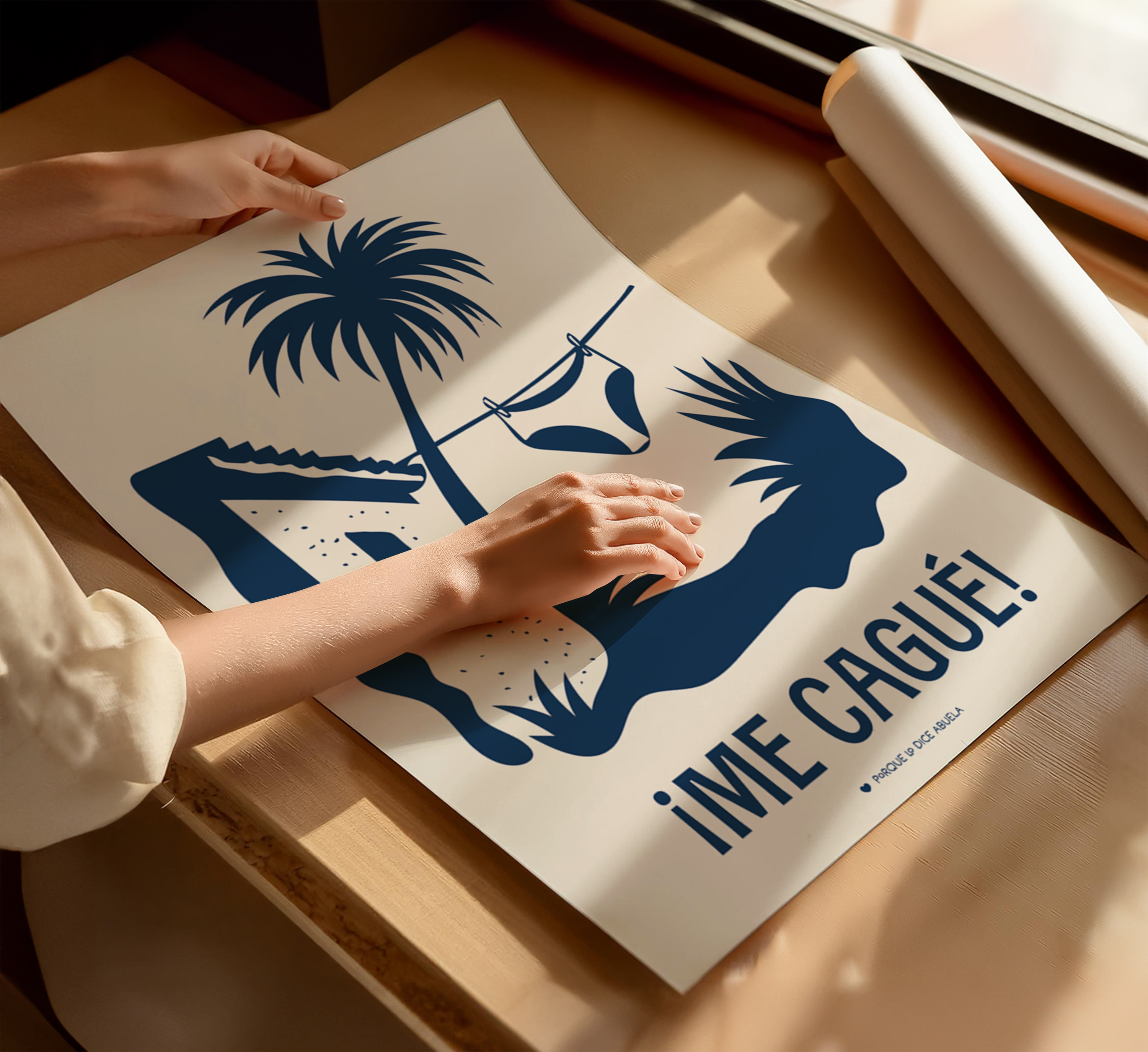 A person's hands are holding a poster with a palm tree and a bikini, along with the text "ime cague!" which translates to "I love Cague!" The poster is placed on a wooden surface, and there is a roll of paper nearby.