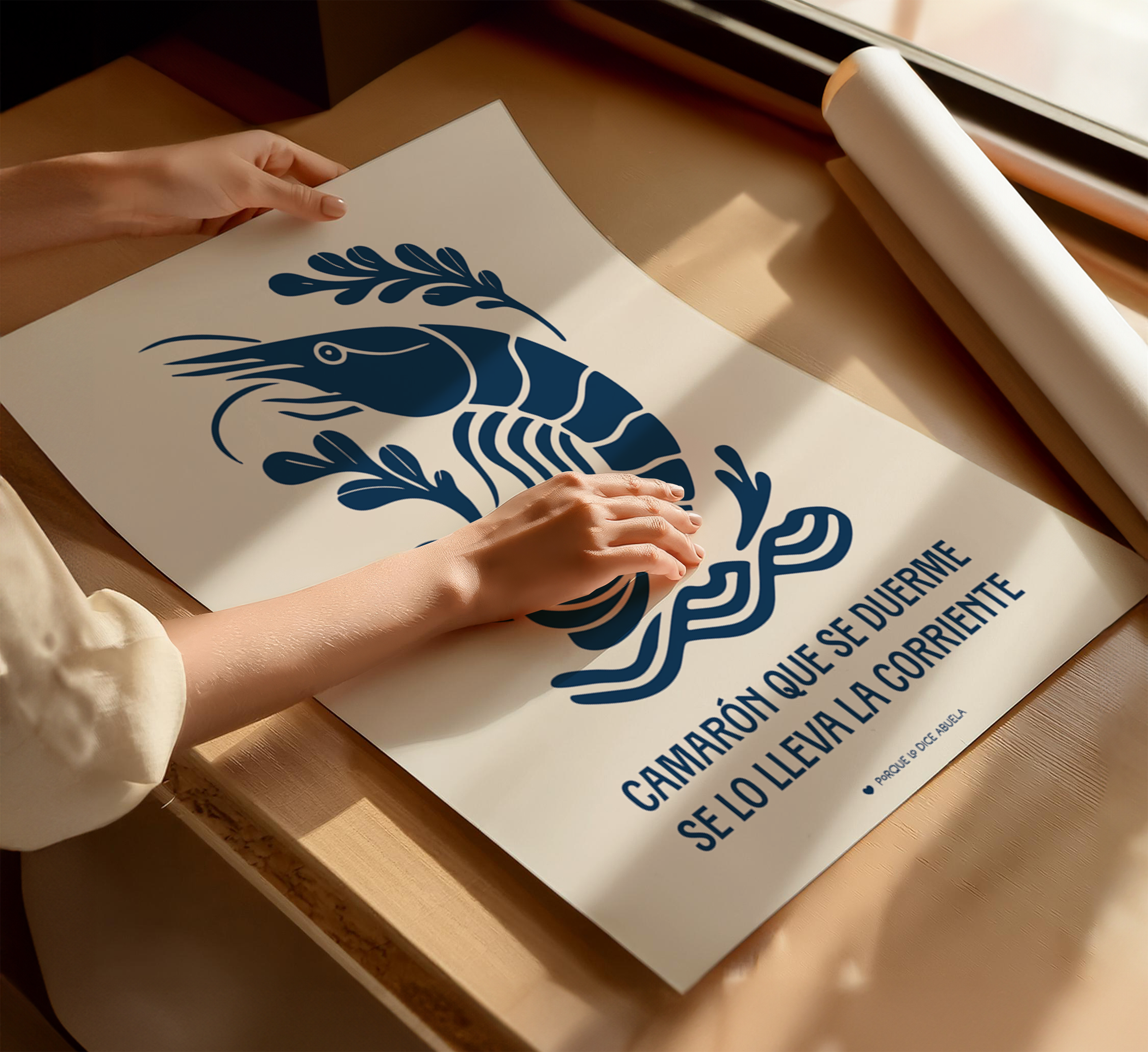 A person's hands are holding a poster with a blue and white design featuring a shrimp and a leafy plant. The poster has text that reads "Camarón que se duerme se lo lleva la corriente" which translates to "A shrimp that swims in the sea".